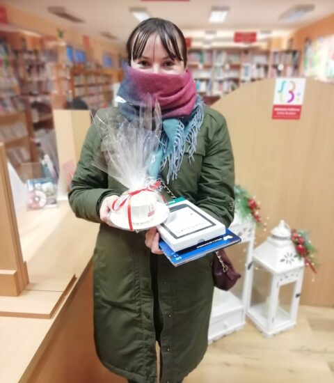na zdjęciu kobieta z kolorową chustą zasłaniającą nos i usta. W rękach trzyma filiżankę owiniętą w folię oraz książkę i czytnik ebooków. W tle jasne meble biblioteczne.