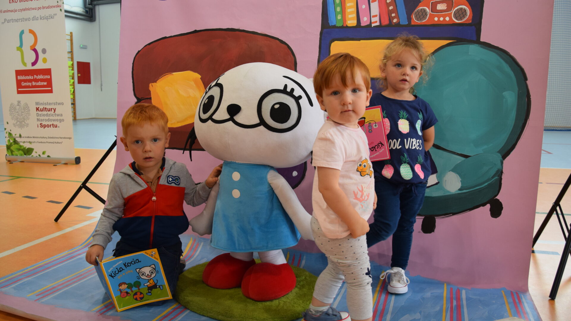 chłopiec trzymający książkę, duża maskatka Kici Koci i dwie dziewczynki na tle ścianki imitującej pokój