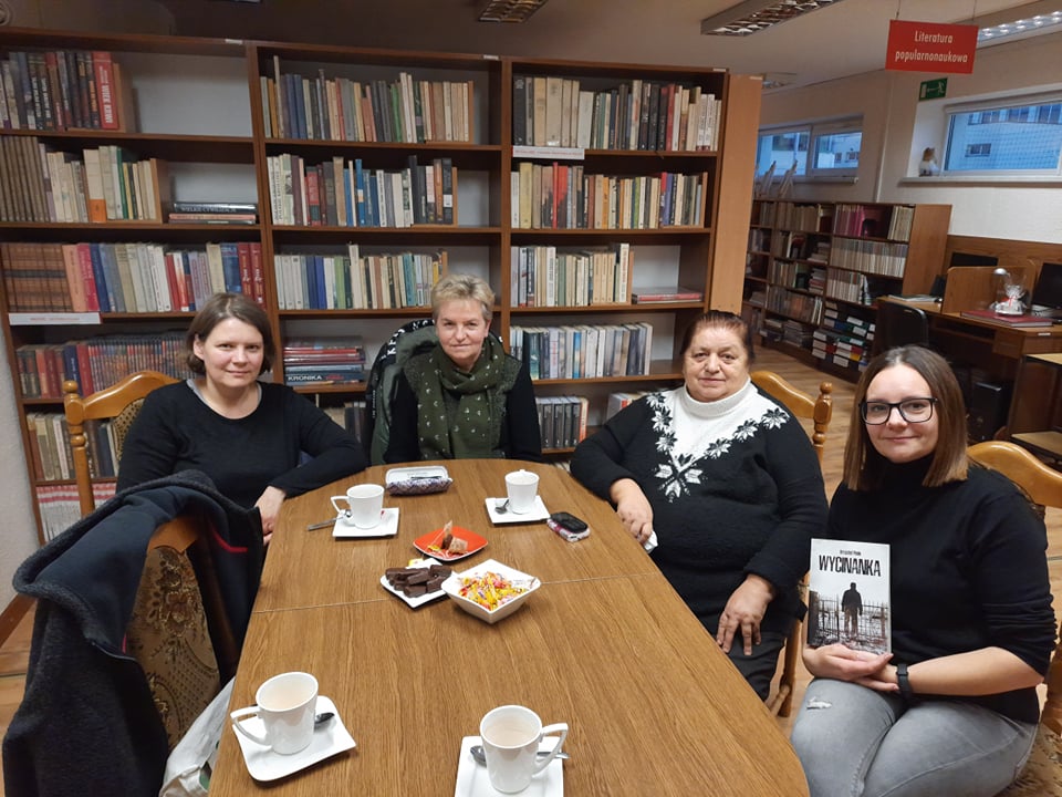 cztery kobiety siedzące przy owalnym stole, jedna z nich trzyma w rękach książkę
