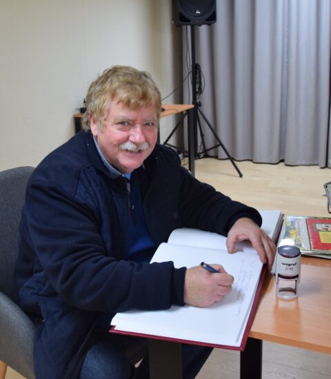 Uśmiechnięty mężczyzna siedzi przy stole przy otwartej kronice, w ręku trzyma długopis