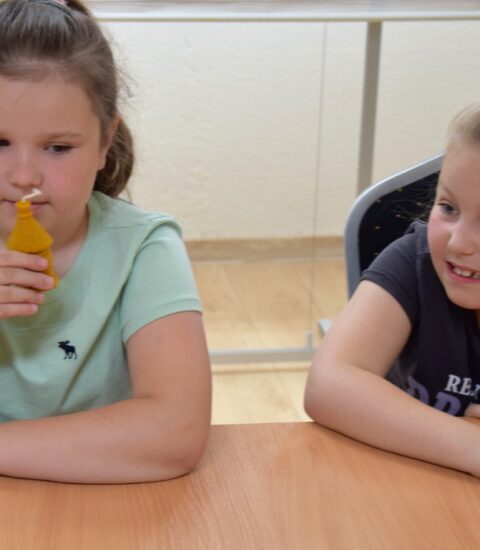 dwie dziewczynki siedzą przy stole, jedna z nich trzyma w rękach figurkę z wosku