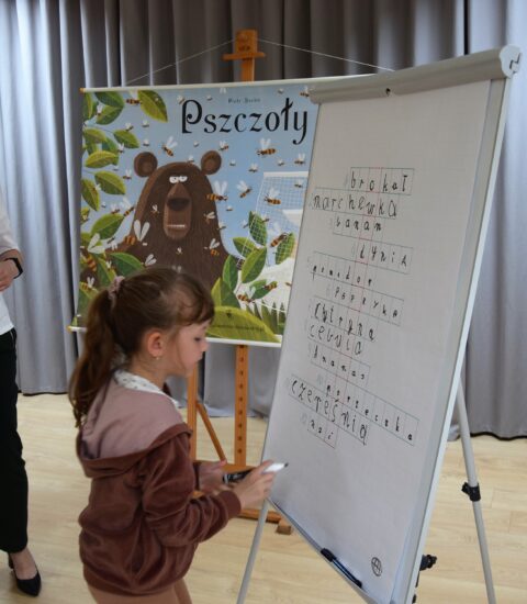 kobieta ubrana w białą bluzkę i czarne spodnie, obok dziewczynka pisze na białej tablicy