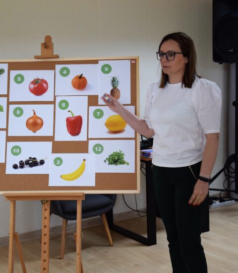 kobieta stoi obok tablicy, na której powieszone są zdjęcia warzyw i owoców, wskazuje ręką