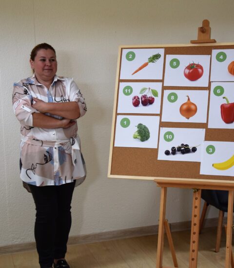 kobieta stoi przy tabliny, na której powieszone są zdjęcia warzyw i owoców