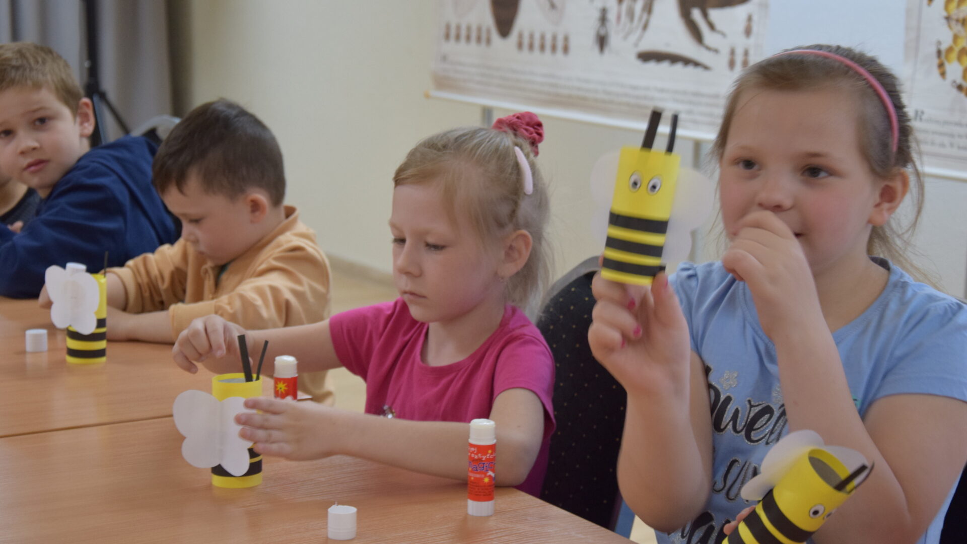 dwie dziewczynki i dwóch chłopców siedzą przy stole, kleją papierowe pszczoły