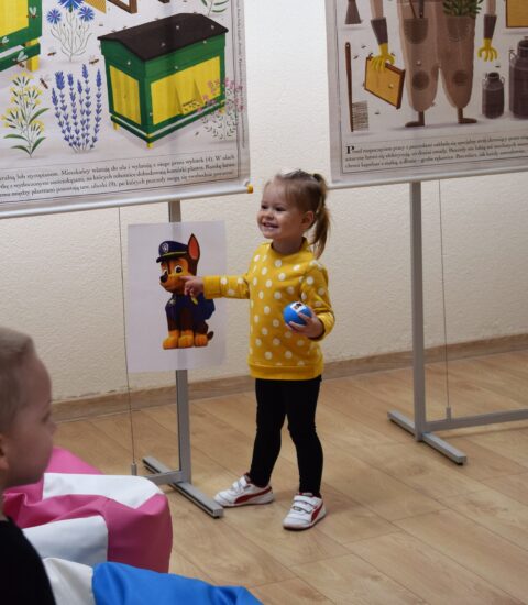 dziewczynka w żółtej bluzce wskazuje palcem obrazek powieszony na stojaku