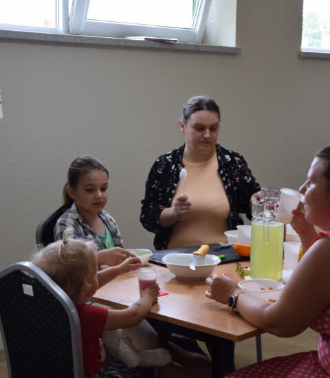 dwie kobiety siedzą przy stole razem z dziećmi, trzecia stoi obok, w ręku trzyma kubek