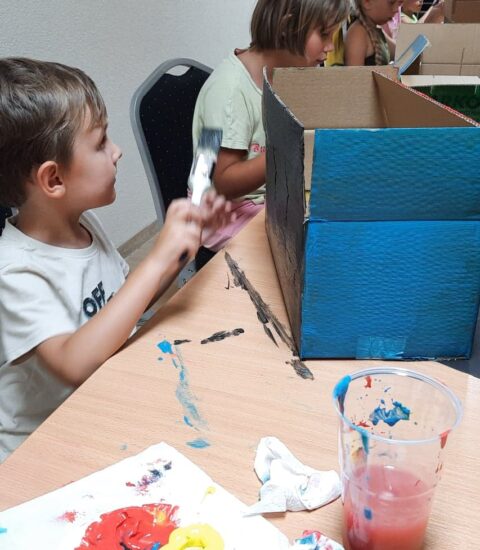 dzieci siedzą przy stołach, malują farbami kartony