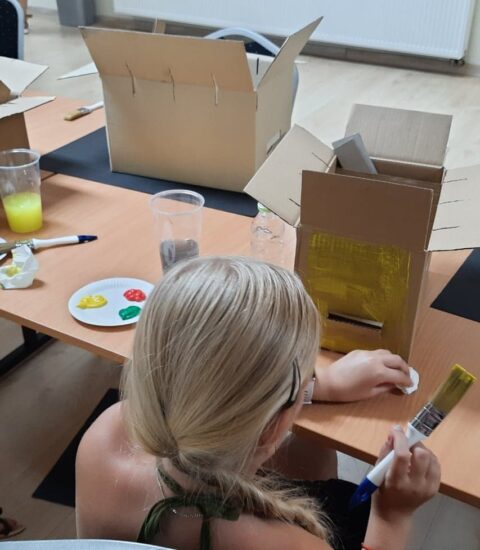 dzieci siedzą przy stole, pędzlami malują kartonowe pudełka