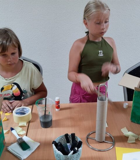 dwie dziewczynki, jedna siedzi przy stole, druga stoi obok, na stole kartony pomalowane farbami