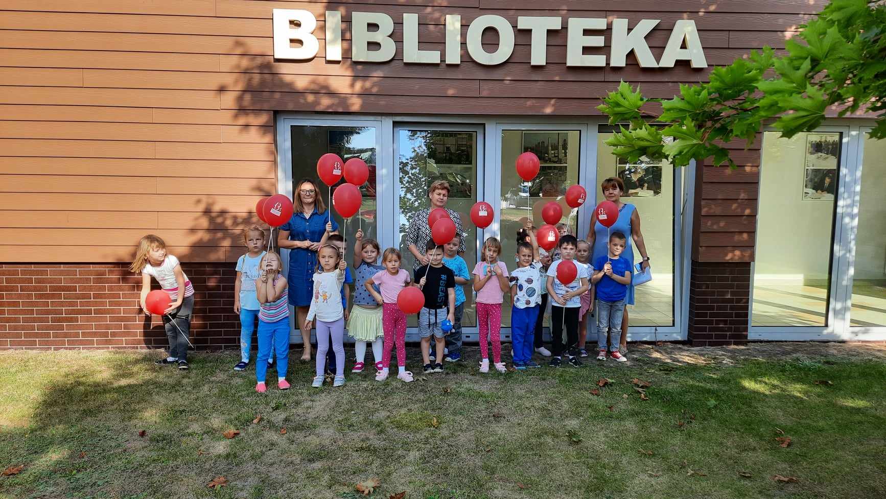 grupowe zdjęcie dzieci i trzech kobiet przed budynkiem biblioteki, dzieci trzymają czerwone balony