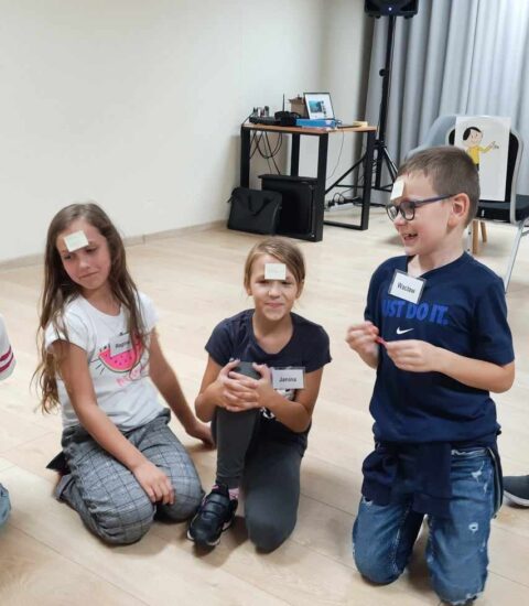 kilkoro dzieci siedzi na podłodze, mają przyklejone kartki do czoła, jeden chłopiec podniesiony na kolanach, wszyscy się uśmiechają