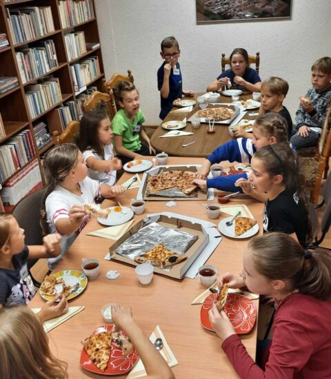 kilkanaścioro dzieci siedzi przy stole, jedzą pizzę