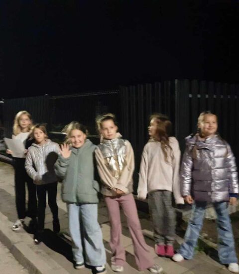 sześć dziewczynek stoi przy ciemnym płocie, niektóre z nich ubrane w kurtki, zdjęcie zrobione nocą