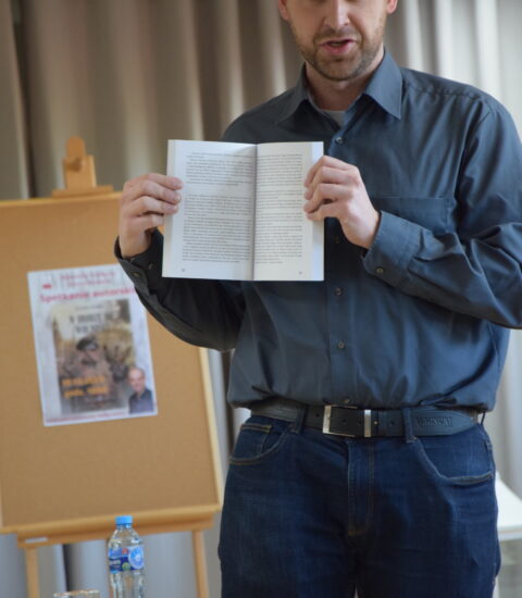 mężczyzna w ciemnej koszuli trzyma w rękach otwarta książkę, skierowaną do obiektywu