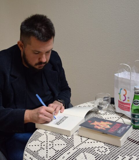 mężczyzna siedzi przy stoliku nakrytym serwetą, pisze długopisem w książce