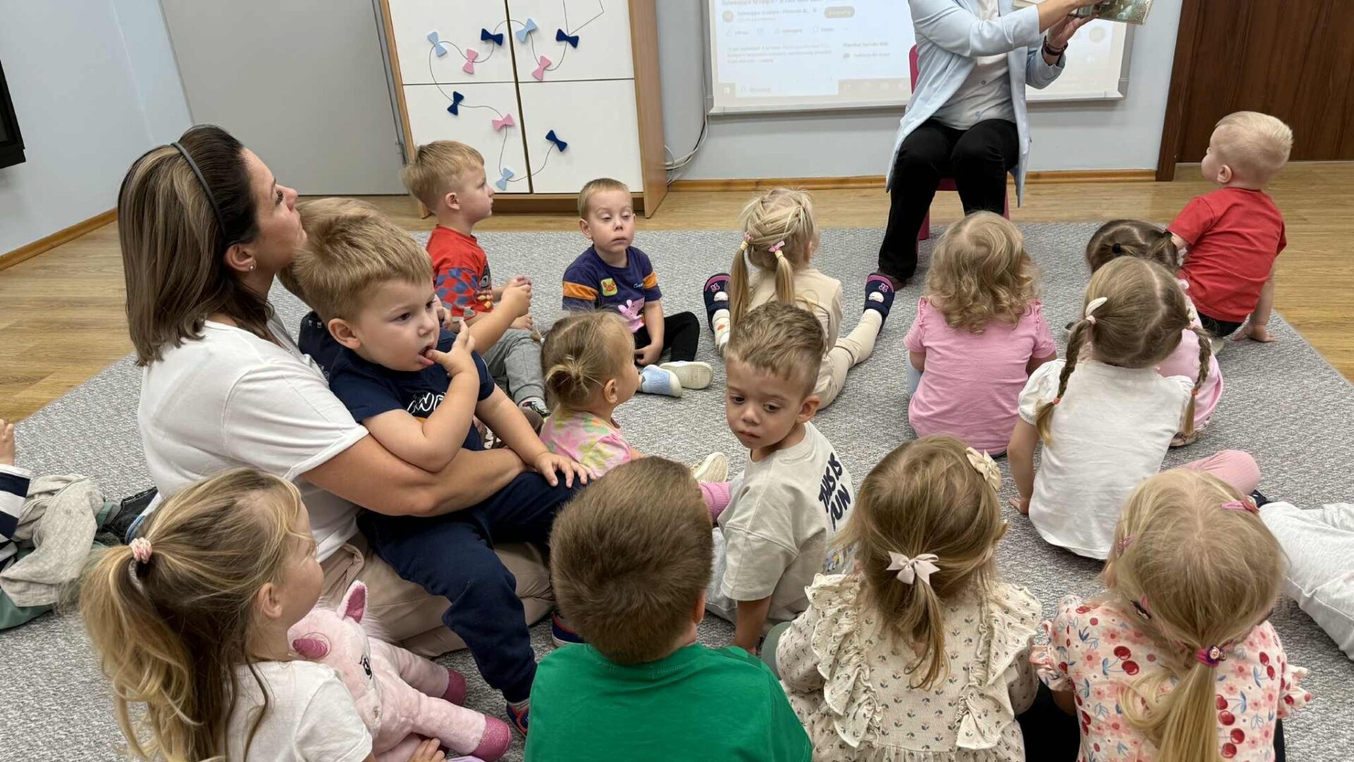 Ogólnopolski Dzień Głośnego Czytania kobieta siedząca na krzesełku pokazuje dzieciom książkę, dzieci siedzą na dywanie