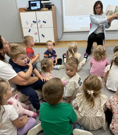 Ogólnopolski Dzień Głośnego Czytania kobieta siedząca na krzesełku pokazuje dzieciom książkę, dzieci siedzą na dywanie