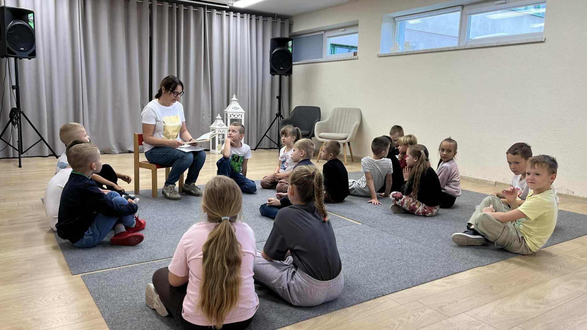 Świętowania ciąg dalszy kobieta siedząca na krzesełku czyta dzieciom książkę, dzieci siedzą na dywanie