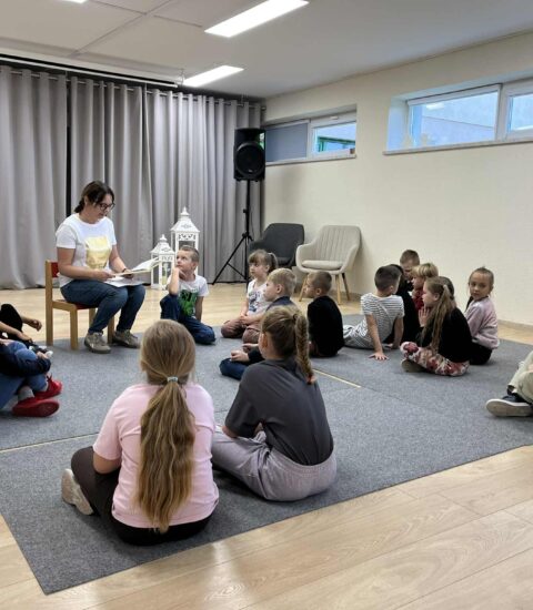 Świętowania ciąg dalszy kobieta siedząca na krzesełku czyta dzieciom książkę, dzieci siedzą na dywanie