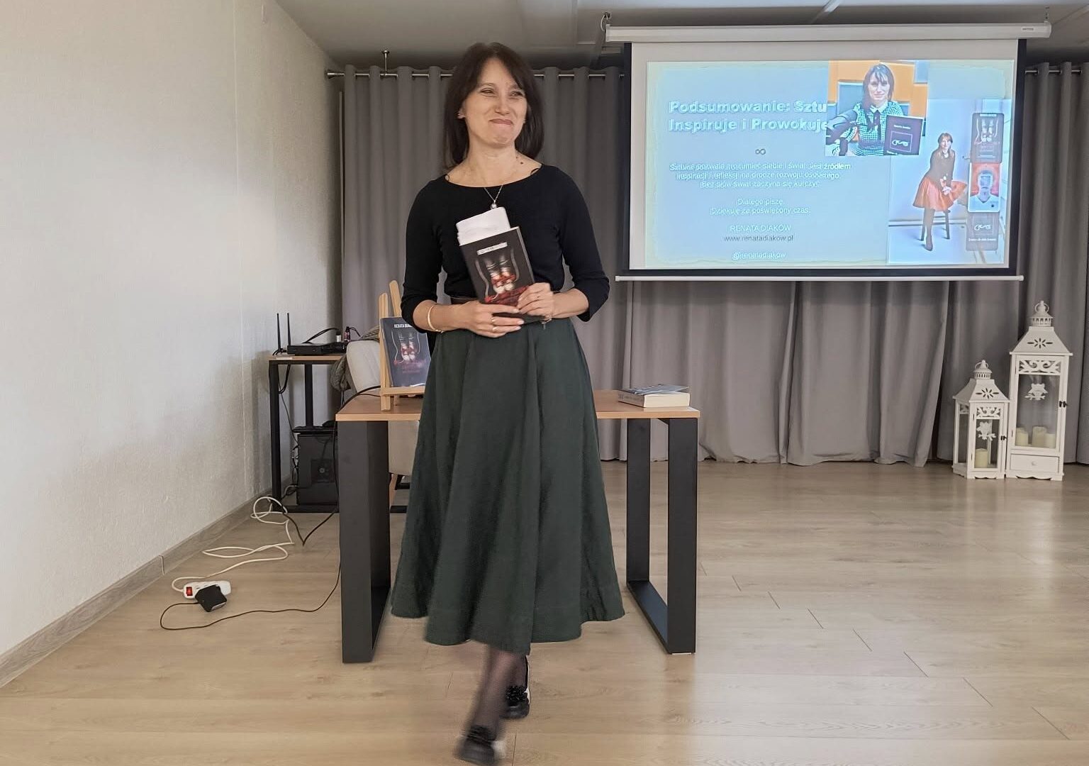 Spotkanie z Renatą Diaków uśmiechnięta kobieta stoi przed ekranem, w rękach trzyma ksiązkę