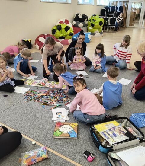 grupa dzieci, razem z rodzicami, siedzą na dywanie, kredki rozłożone, dzieci kolorują