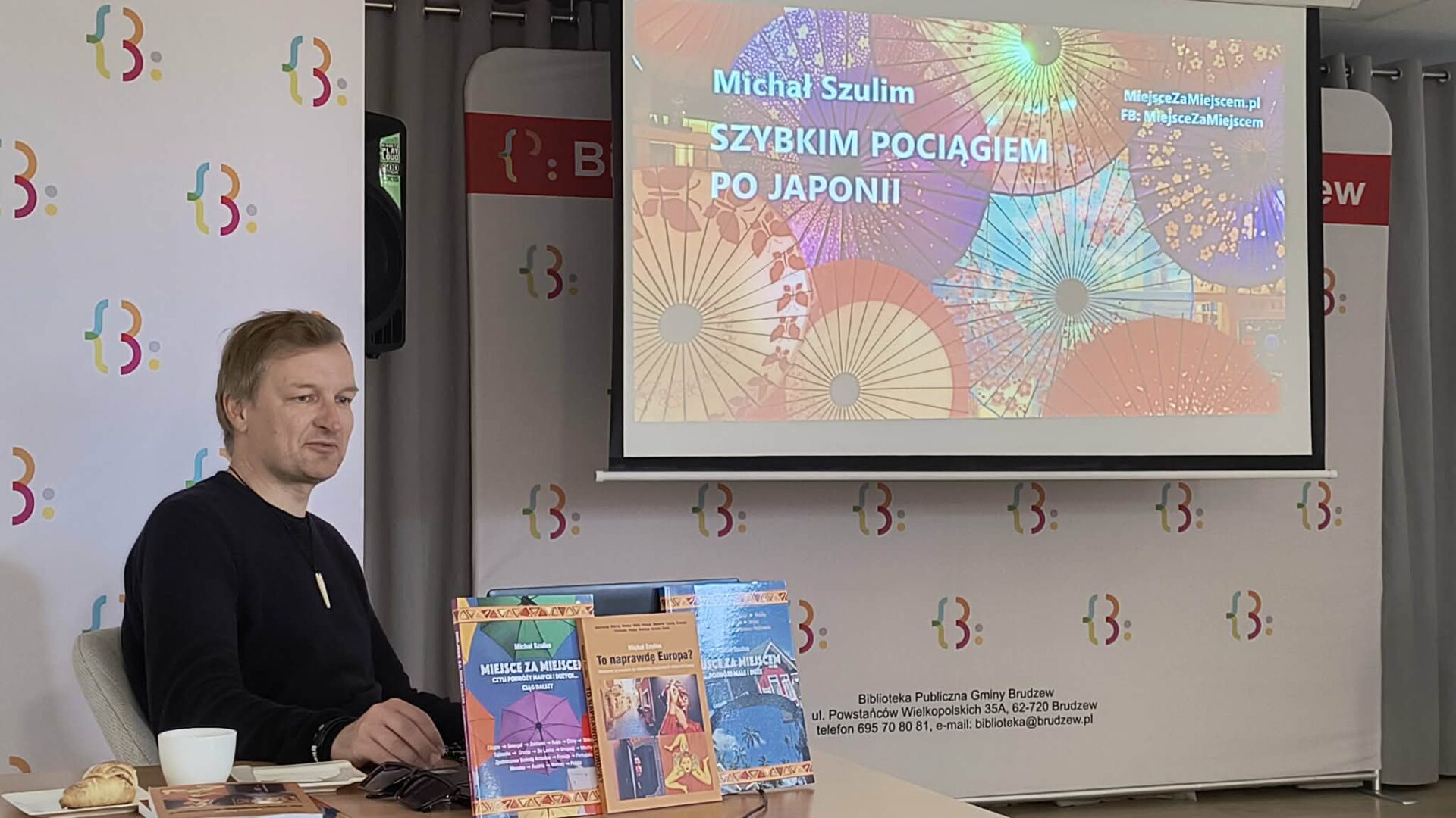 mężczyzna siedzi przy stoliku, w tle ekran, na ktorym wyświetlany jest kolorowy obraz