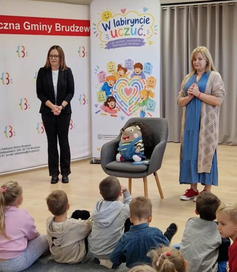 dyrektor biblioteki i autorka stoją przed dziećmi, pomiędzy nimi na fotelu siedzi pluszowy jeż